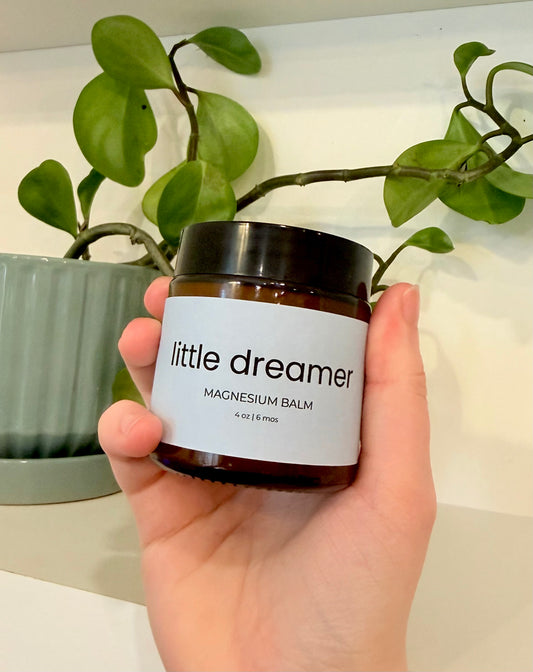Hand holding a jar of 'little dreamer' magnesium balm with a plant in the background.
