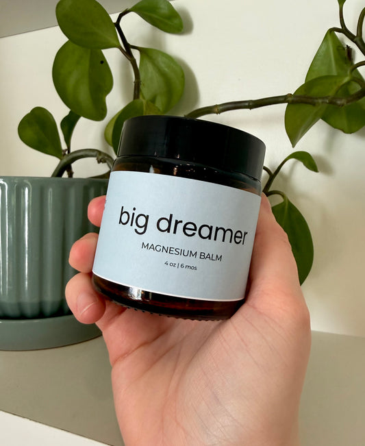 Hand holding a jar of 'big dreamer' magnesium balm with a plant in the background