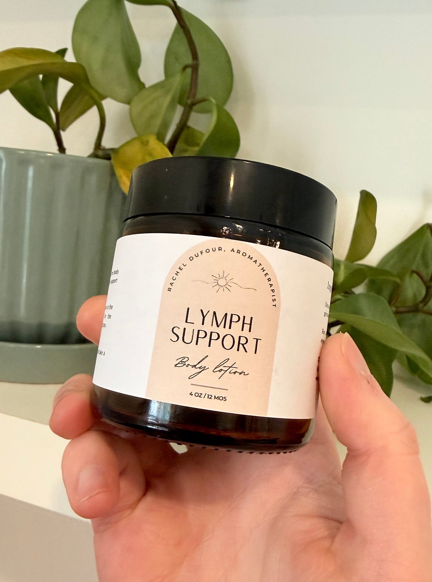 hand holding a jar of body lotion labeled "lymph support" with a plant in the background