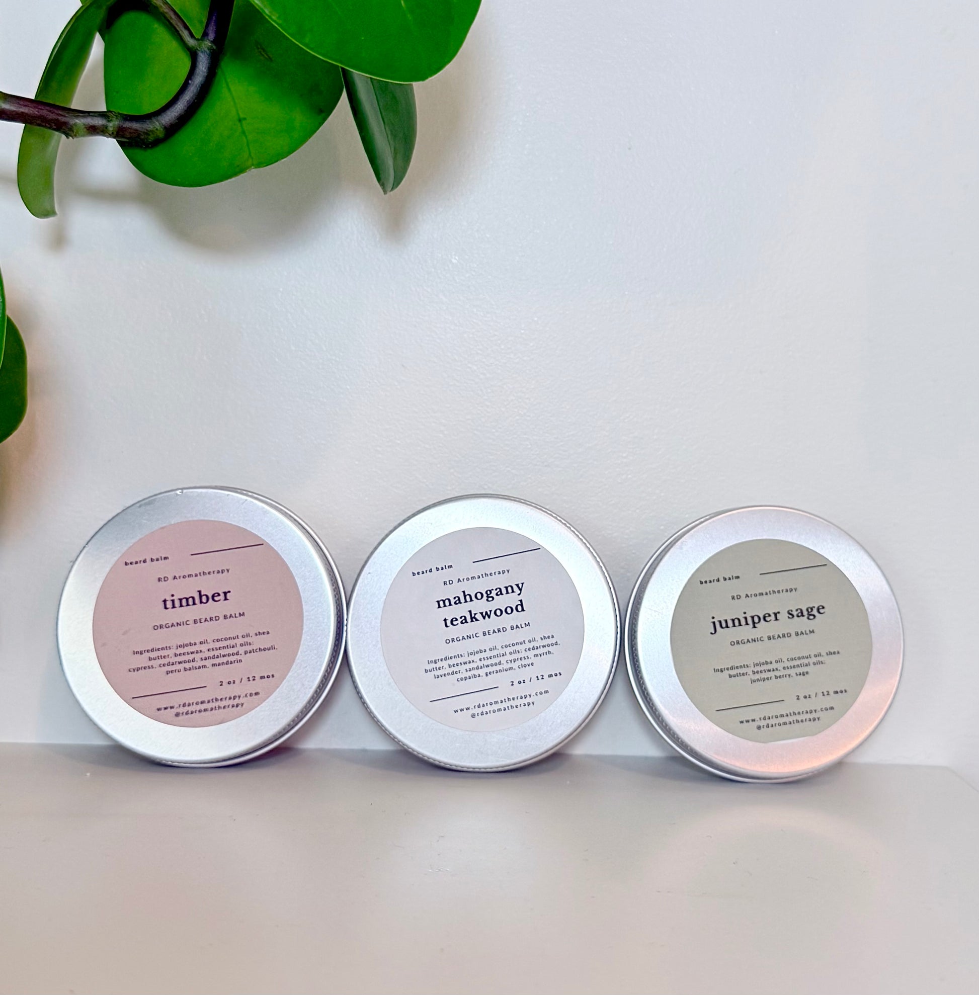Three tins labeled 'timber', 'mahogany teakwood', and 'juniper sage' on a light surface with green leaves in the corner.