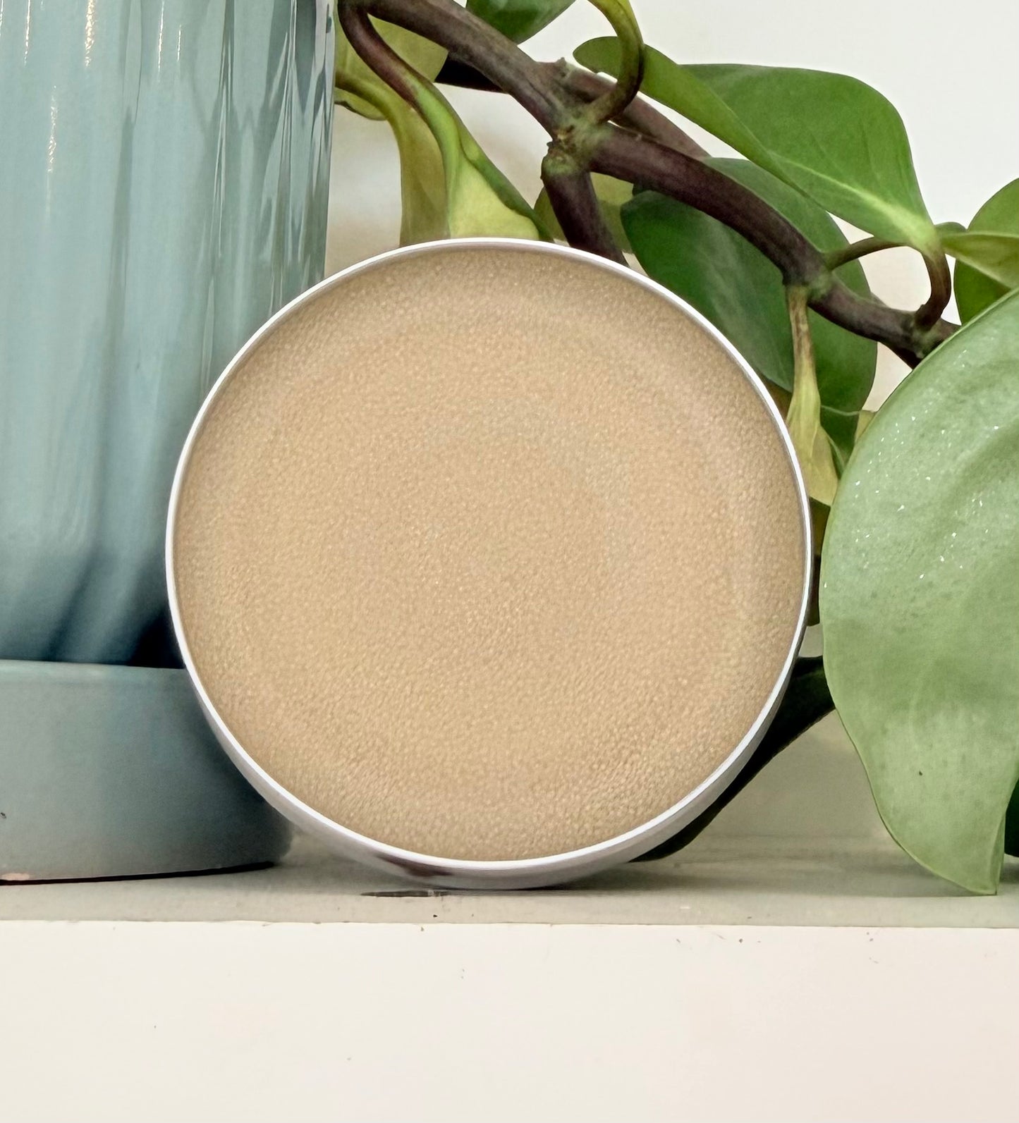 Round beige powder in a metal container with green plants in the background
