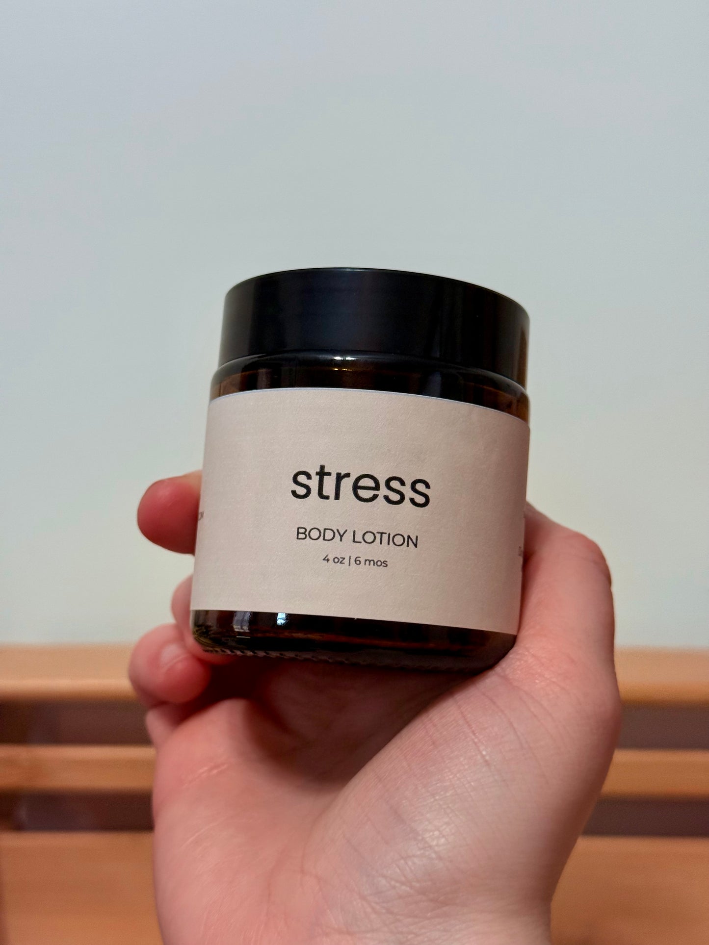 Hand holding a jar of body lotion labeled 'stress' against a neutral background