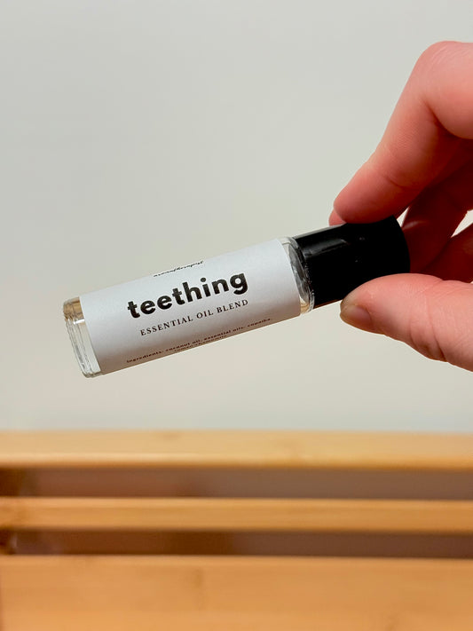 Hand holding a bottle labeled 'teething essential oil blend' against a neutral background