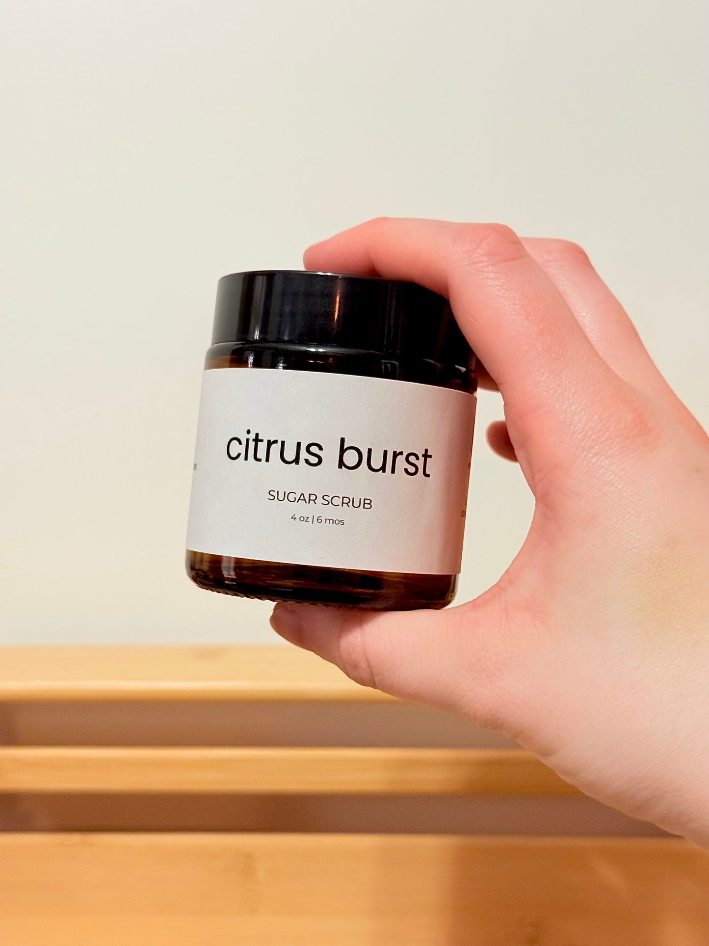 Hand holding a jar of 'citrus burst' sugar scrub against a neutral background