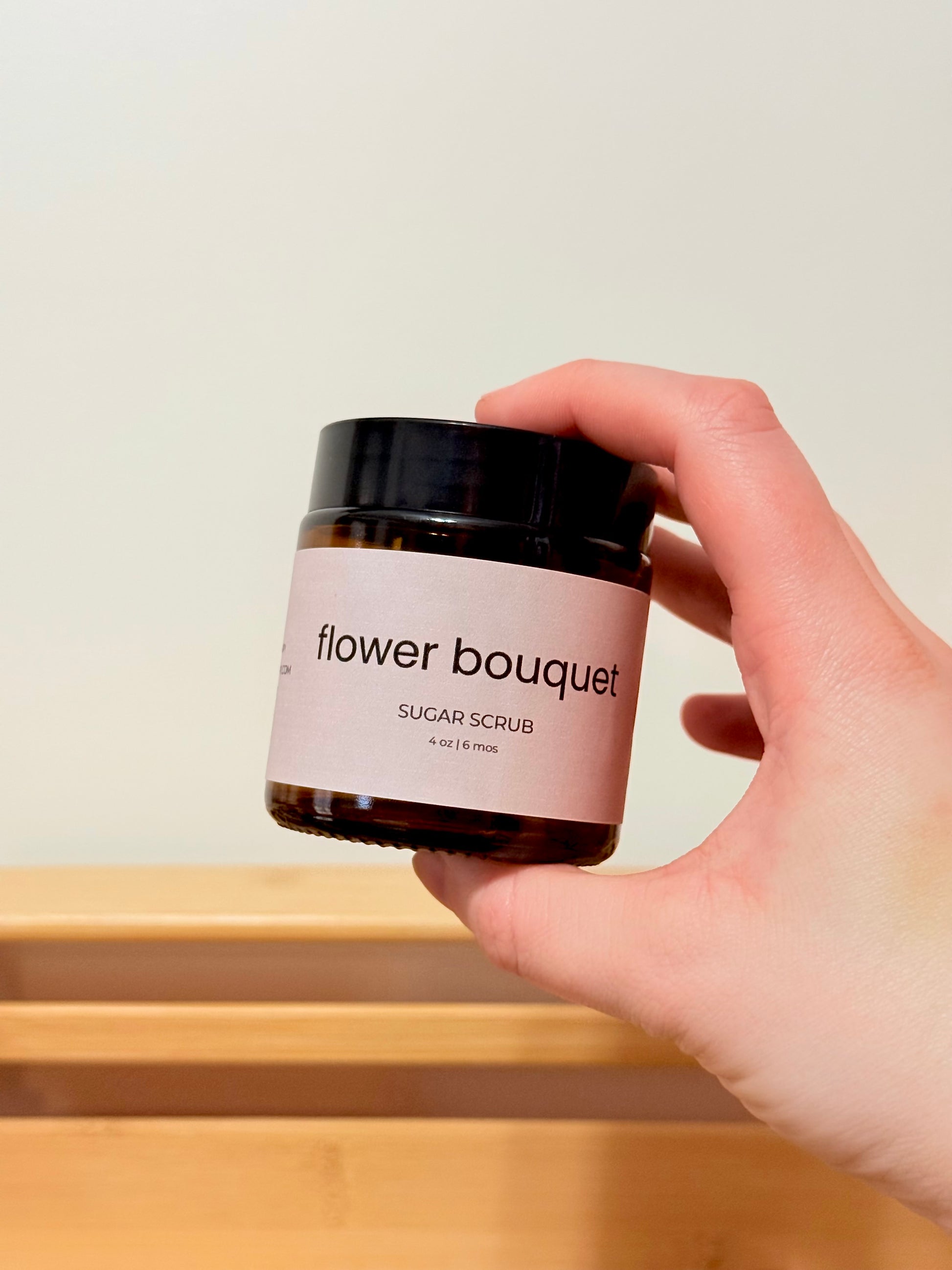 Hand holding a jar of 'flower bouquet' sugar scrub against a neutral background