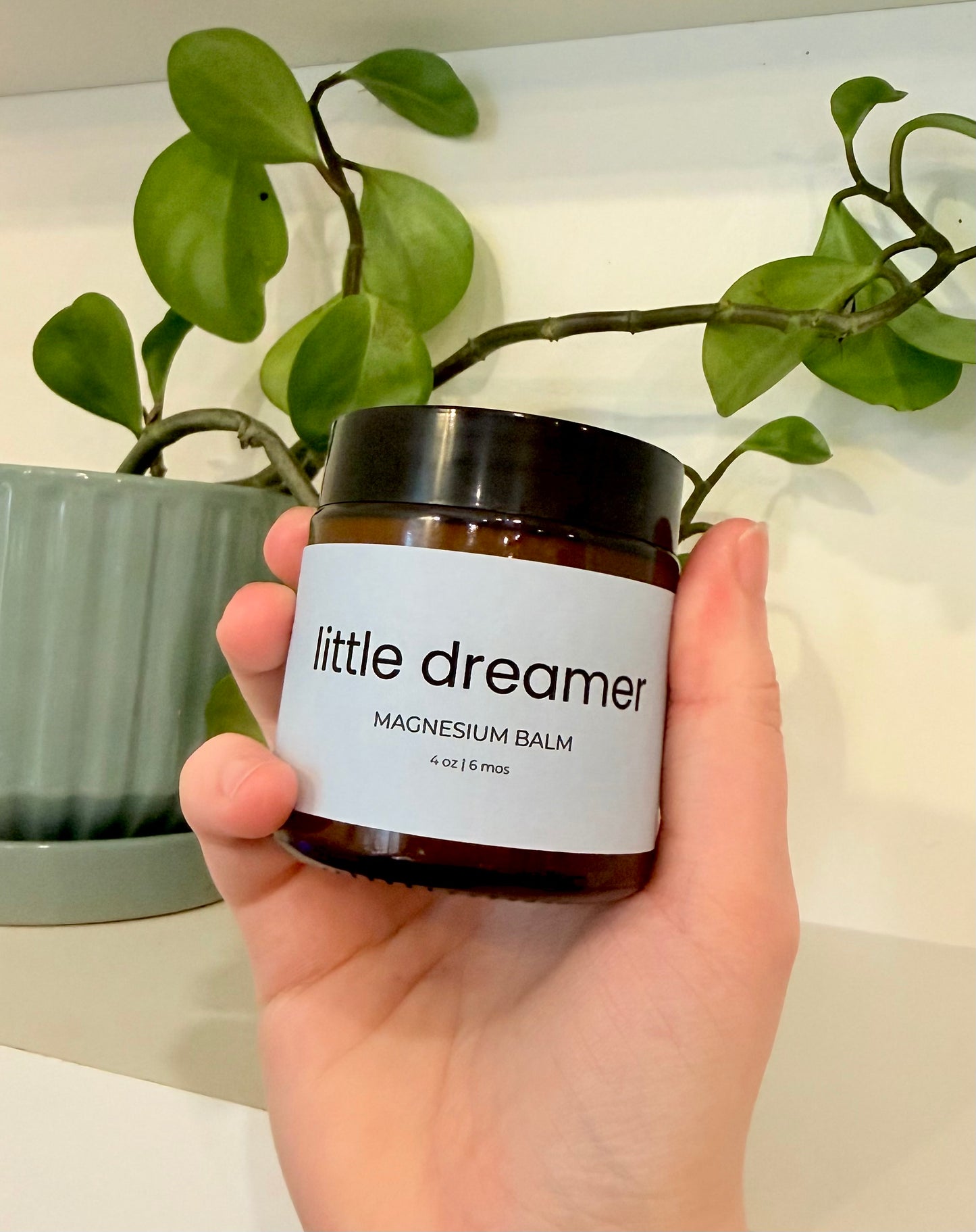 Hand holding a jar of 'little dreamer' magnesium balm with a plant in the background.
