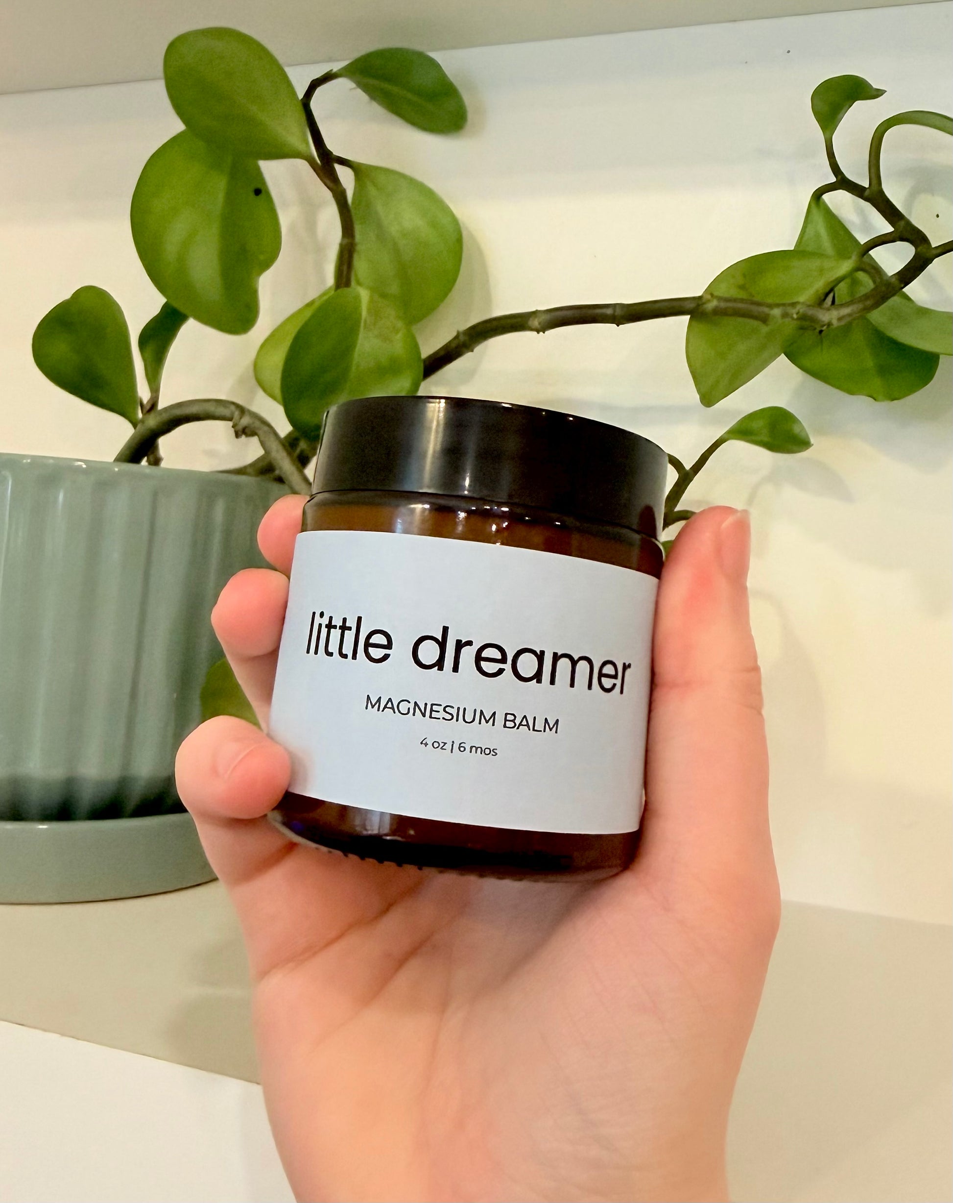 Hand holding a jar of 'little dreamer' magnesium balm with a plant in the background.