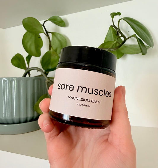 Hand holding a jar of 'sore muscles' magnesium balm with plants in the background